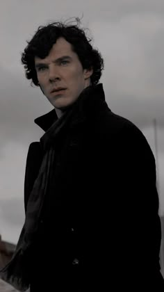 a man with curly hair wearing a black coat and standing in front of a cloudy sky