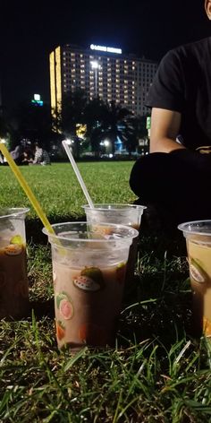 four cups with drinks in them sitting on the grass near a man holding a cell phone