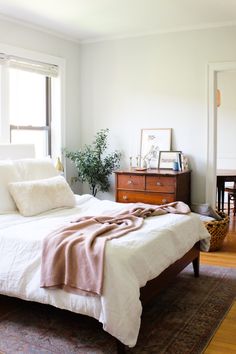 a bed with white sheets and pillows in a bedroom next to a dresser, chair and window