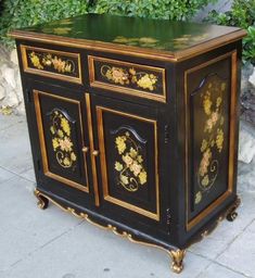 an antique black and gold painted cabinet with flowers on the front, sitting on a sidewalk