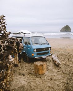 a blue van parked on top of a beach next to a tree stump and surfboard