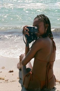 a woman sitting on top of a beach holding a camera next to the ocean while wearing a bathing suit