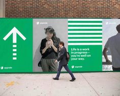 a man walking past a green sign with an arrow on it's side and a woman taking a photo