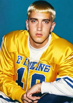 a young man with blonde hair wearing a yellow jacket and white shirt sitting on a blue background