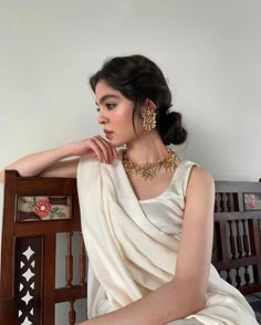 a woman sitting on top of a wooden chair wearing a white dress and gold jewelry