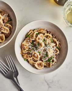 two bowls filled with pasta and meat on top of a white table next to silverware