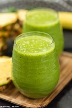 two glasses filled with green smoothie sitting on top of a cutting board