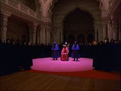 two women in purple dresses stand on a pink carpet with other people standing behind them