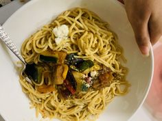 a white bowl filled with pasta and vegetables on top of a table next to a person's hand