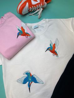 two t - shirts with colorful birds on them next to a pair of tennis shoes