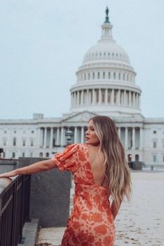 a woman in an orange dress leaning on a railing with the capital building in the background