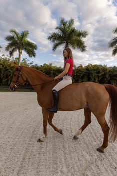 a woman riding on the back of a brown horse across a sandy field next to palm trees