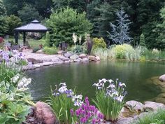 a pond surrounded by rocks and flowers with a gazebo in the background