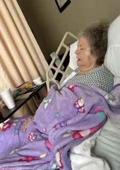 an old woman laying in a hospital bed with a purple blanket over her head and she is looking at the camera