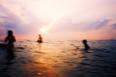 three people are in the water on surfboards at sunset or dawn, with one person standing up and another sitting down