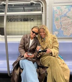 two women are sitting on a subway car and one is holding a camera while the other looks at her phone