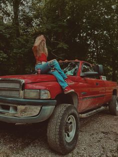 a woman sitting on the hood of a red pickup truck in front of some trees