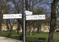 two street signs on a metal pole in front of some benches and trees with buildings in the background