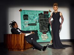 a woman in a black dress standing next to a man sitting on a desk with a painting behind her