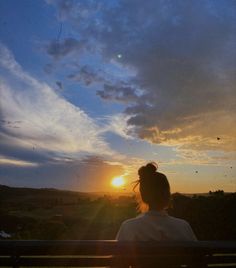 a woman is sitting on a bench watching the sun go down with her hair blowing in the wind