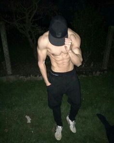 a shirtless man in black pants and a hat standing on the grass at night