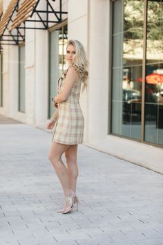 a woman standing on the sidewalk wearing a tan dress and high heeled sandals with her hand in her pocket