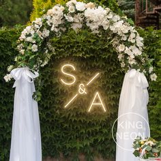 the wedding arch is decorated with white flowers and greenery as well as an illuminated s & a sign