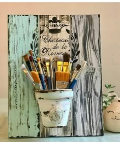 a cup filled with lots of paint brushes sitting on top of a wooden sign next to a