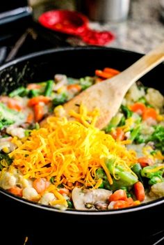 a skillet filled with vegetables and cheese on top of a stove burner next to a wooden spoon