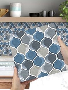 a person holding up a blue and gray tile pattern on the wall next to a bookshelf