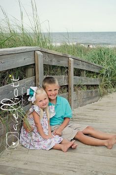 two young children sitting on a wooden walkway near the beach with grass and sea oats in the background