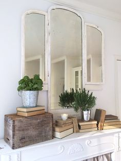 a white mantle with books, plants and mirrors on it in a room that is painted white