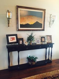 a black console table with pictures and plants on it in front of a framed painting