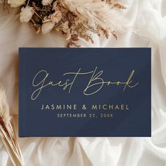 a blue guest book with gold lettering on it next to some dried flowers and feathers