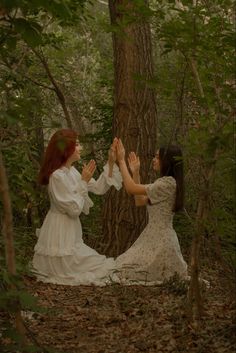 two women in white dresses holding hands up to each other while standing next to a tree