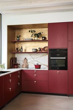 a kitchen with red cabinetry and white counter tops is pictured in this image, there are plants on the shelves above the sink