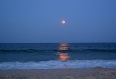 the moon is setting over the ocean with waves coming in to shore and blue sky