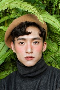 a young man wearing a brown hat standing in front of green plants and ferns, looking at the camera