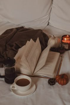 an open book and cup of coffee sit on a bed next to a blanket, candle, and pumpkins