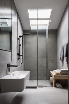 Modern bathroom with a skylight, glass shower, wall-mounted sink, and wooden bench with folded towels.