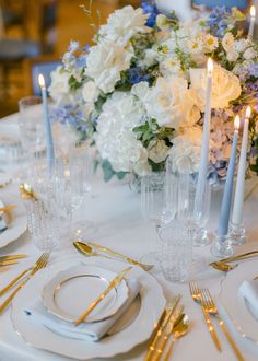 the table is set with white and blue flowers, silverware, and gold place settings