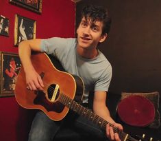 a young man playing an acoustic guitar in a room with red walls and pictures on the wall