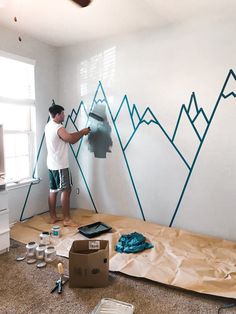 a man is painting a mountain mural on the wall in his living room with blue tape