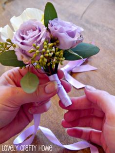 two hands are holding a bouquet of purple roses and greenery, with the ribbon tied around it