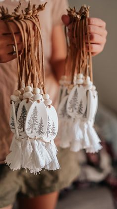 two white ornaments hanging from strings with pine trees on them and tassels in the air