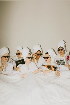 four women in white robes and sunglasses are sitting on a bed with books, reading