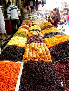 many different types of fruits and vegetables on display at an open air market with people standing around