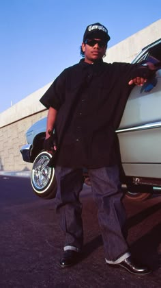 a man standing in front of a car with his hand on the wheel and wearing sunglasses