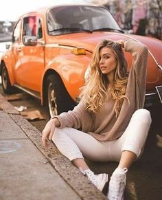 a woman is sitting on the curb next to an orange car and posing for a photo