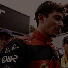 a man standing next to another man wearing a red and black racing suit on top of a race track